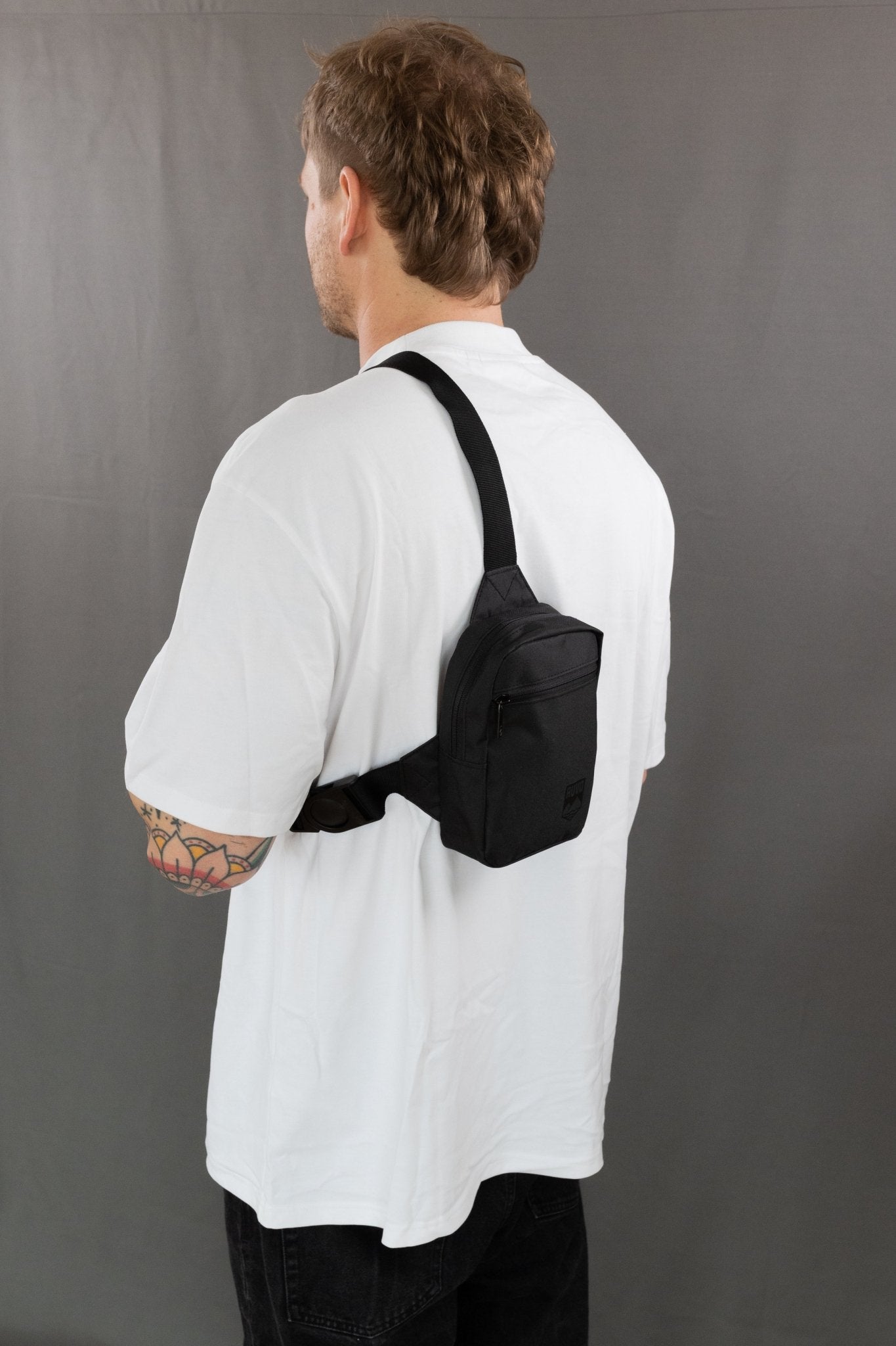 RUVO Chest Bag Größe S in der Farbe schwarz mit einem männlichen Modell. Dieses hat ein weißes T-Shirt an und steht vor einer grauen Wand.