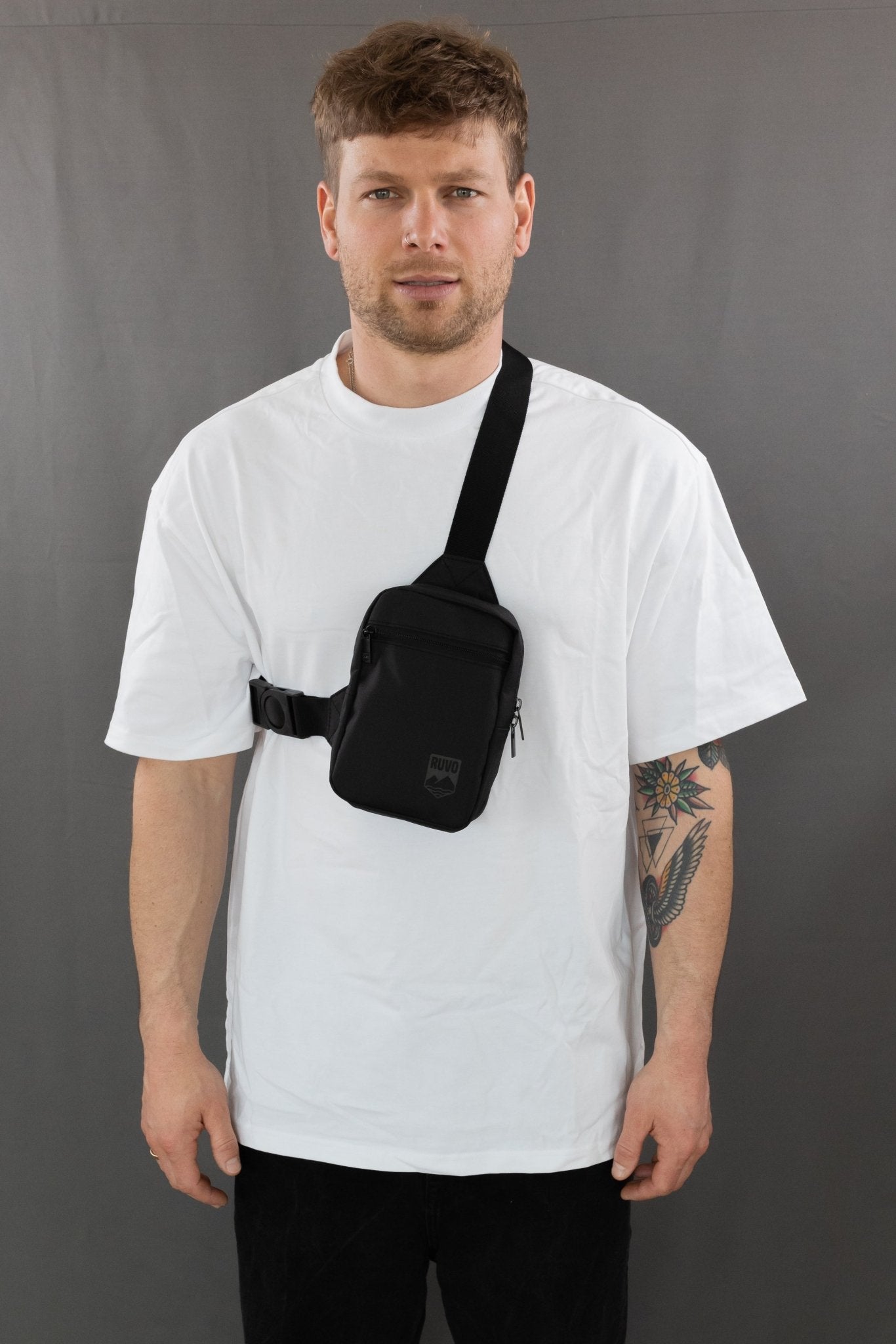 RUVO Chest Bag Größe S in der Farbe schwarz mit einem männlichen Modell. Dieses hat ein weißes T-Shirt an und steht vor einer grauen Wand.