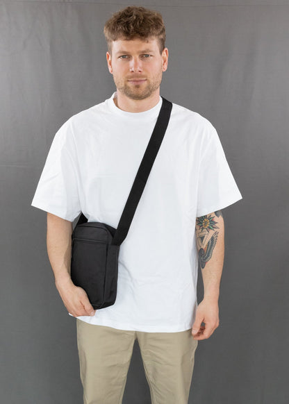 RUVO Umhängetasche Sling Bag Größe M in schwarz. Modell trägt die Tasche über der linken Schulter und man sieht das Modell von vorne. Die Hand des Modells liegt an der Tasche an.